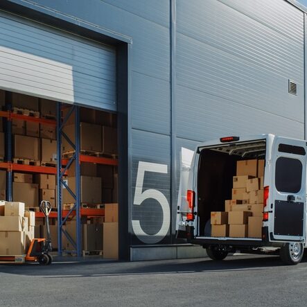 Outside of Logistics Warehouse with Open Door, Delivery Van Loaded with Cardboard Boxes. Truck Delivering Online Orders, Purchases, E-Commerce Goods, Wholesale Merchandise.